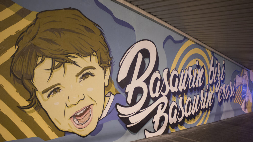 Mural en el que se ve un niño riendo junto a las letras: Basaurin bizi, basaurin erosi