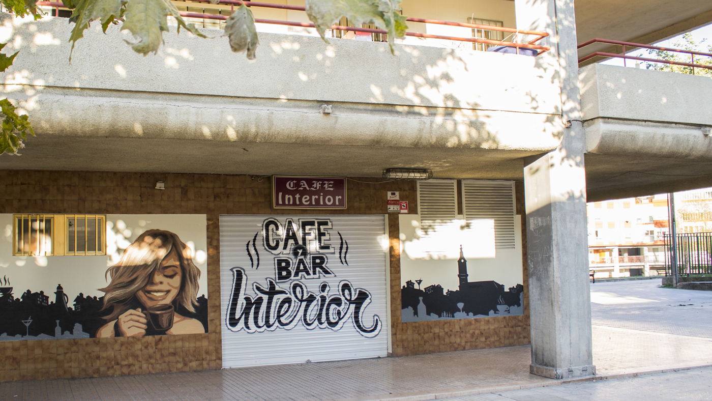 Mural realizado por Ha13estudio en el exterior del bar Interior, en el mural se ve una chica tomando café, y la rotulación sobre la persiana del nombre del bar.